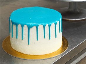A white frosted cake with vibrant blue dripping icing on a stainless steel surface ready to be