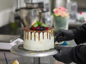 Decorating a cake with a golden drip, green leaves and fresh berries on a cake stand in a kitchen