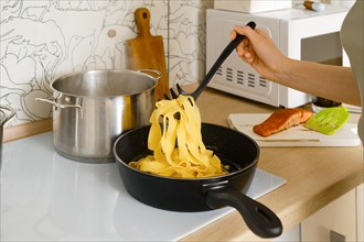 A woman transfers cooked pasta into a frying pan with fried vegetables