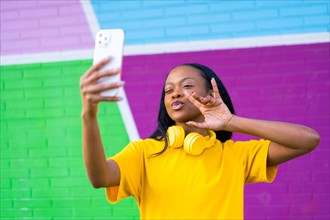 African cool woman gesturing with headphones on the neck taking a selfie against a colorful wall