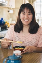 Vertical portrait of Asian woman sitting at home eating rice with chopsticks looks and smiles at