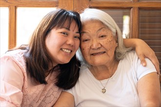 Close up of Japanese adult daughter hugging her grey haired mother smiling and looking at the