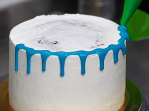 A white frosted cake in the process of being decorated with blue icing with a filling bag with