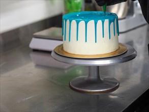 A frosted cake with white frosting being decorated with blue icing on a kitchen counter by pastry
