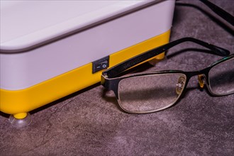 Closeup of pair of wire framed glasses in front of ultrasonic cleaner machine on black textured