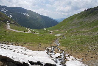 A winding road meanders through a green valley with snowfields, Capra River, mountain road,