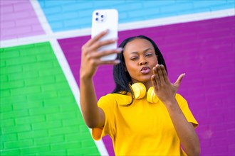 African woman blowing a kiss while taking a selfie against a colorful urban wall outdoors
