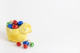 A yellow ceramic chick filled with a selection of colourful chocolate eggs, white background, copy