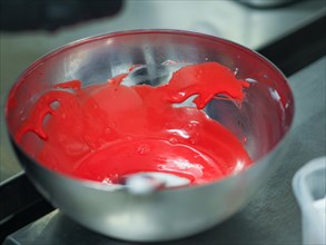 Shiny red icing being prepared in a metal bowl in professional kitchen for frosted cake decoration