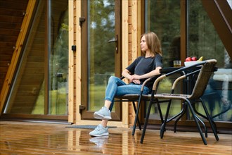 Cute woman enjoys fresh air after the rain sitting on terrace of her forest cabin