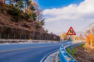 Warning sign next to guardrail on curvy mountain road with cloudy sky in background in South Korea