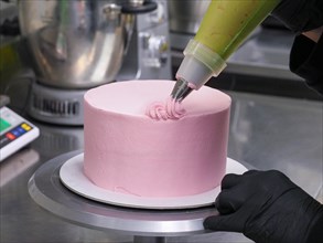 Close-up of a confectioner's piping bag adding pink frosting to decorate a cake with swirls,