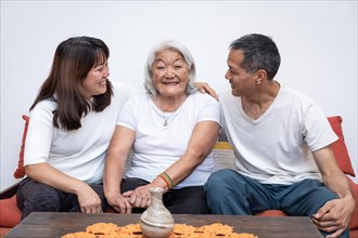 Adult children, with smiles on their faces, embrace their joyful elderly mother as she sits on a