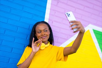Smiling african woman waving while taking a selfie with the mobile phone against a colorful wall