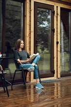 Cute woman in casual clothing sits reading on a wet terrace of a log cabin after the rain, enjoying
