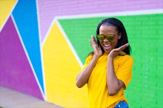 Excited african girl with sunglasses screaming joyful next to a colorful wall in the city