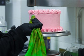 Pink cake being decorated with frosting from a green piping bag on a metal stand in a bakery by