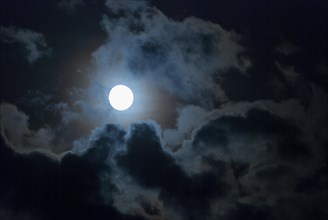 The full moon shines brightly in a cloudy, stormy, black sky and has a halo (corona),