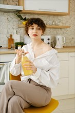 Young woman sits in the kitchen, holds glass bottle of juice in hands and looks to the side