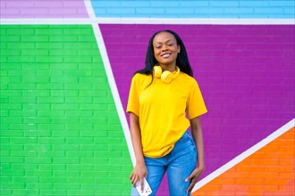 African young woman standing with mobile home and headphones against a colorful wall