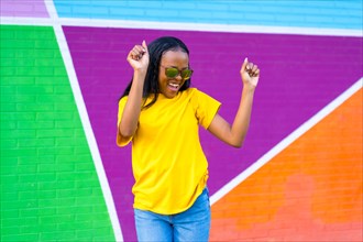 Frontal portrait of a young and happy african woman smiling and dancing next to colorful wall