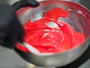 Pasty chef hands wearing black latex gloves stir on A shiny metal bowl full of bright red frosting