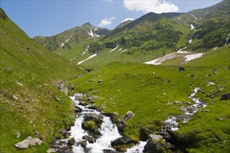 A clear mountain stream flows through green meadows below snow-covered slopes, Capra River,