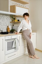 Barefoot young woman standing gracefully in a well-lit modern kitchen. She is wearing white shirt