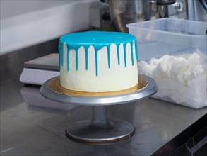 A white frosted cake with vibrant blue dripping icing on a cake stand ready for decoration