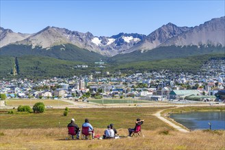 A family sits in chairs in a meadow with a view over the city of Ushuaia and the mountains Cerro