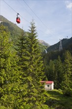 Small cable car gondola crosses a picturesque mountain landscape with fir trees and a small hut and