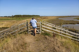 Woman resting at gate on coastal footpath between River Ore and Hollesley Marshes, Suffolk,