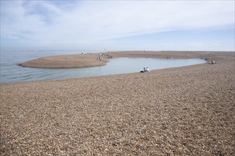 People enjoying a clam day sitting around a recently formed lagoon on the beach at Shingle Street,