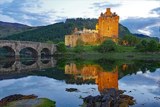 The illuminated Eilean Donan Castle is reflected in the water on a calm evening, old stone bridge,