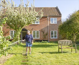 Woman watering garden with hosepipe in front of red brick historic detached house, Shottisham,