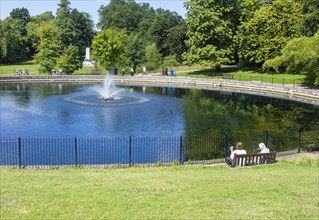 People sitting relaxing on sunny day by water fountain pond in Christchurch Park, Ipswich, Suffolk,