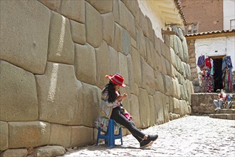 Peruvian woman with hat sitting in front of the traditional Inca wall in Calle Hatunrumiyoc, old