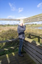 Male birdwatcher ornithologist using binoculars hide at RSPB Hollesley Marshes, Suffolk, England,