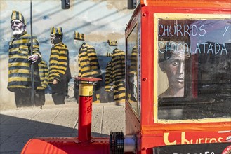 Red trolley for selling churros, behind a graffito with men in convict clothing, Ushuaia, Tierra