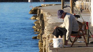 A man sitting on a folding chair fishing on the waterfront in the morning light, Gythio, Mani,