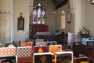 Knitted woollen pew kneelers rest on wooden pews, view towards altar and stained glass east window,