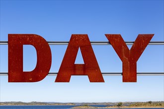 Large red rusty capital letters spell the word DAY against blue sky background, Alqueva dam, Moura,
