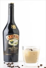 A glass with Baileys and ice, surrounded by coffee beans, a bottle in the background out of focus