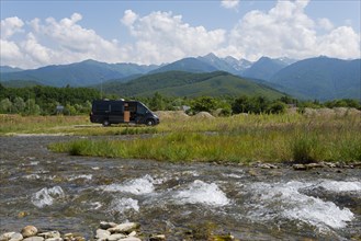 A camper stands on a sunny day next to a river with a view of a mountain range, CarÈ›iÈ™oara River,