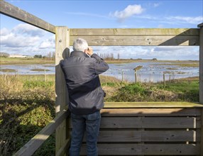 Male birdwatcher ornithologist using binoculars hide at RSPB Hollesley Marshes, Suffolk, England,