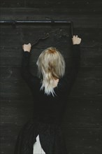 Back view of a blonde thin white woman dressed in black reaching up to a rusty antique metal lock