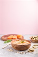 Carrot cream soup with sesame seeds and snacks in wooden bowl on a gray and pink background with