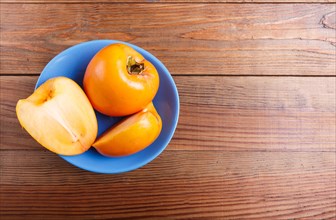Ripe orange persimmon in a blue plate, on brown wooden background, with copy space