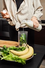 Unrecognizable woman pouring milk into blender with banana and spinach