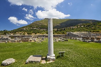Ancient column stands in the foreground of a historical site with green meadow and mountains,